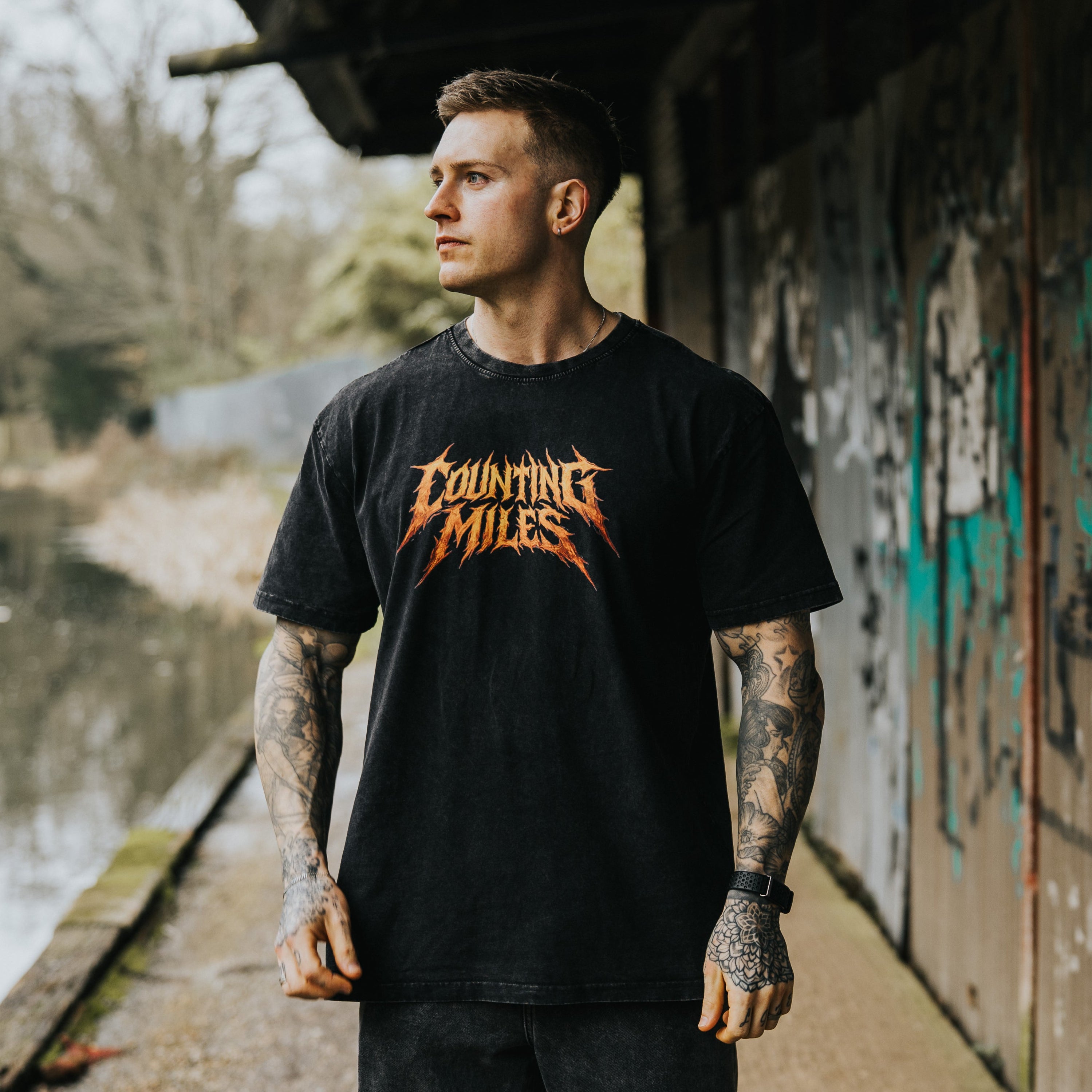 Man wearing a black t-shirt with 'Counting Miles' logo standing near a graffiti-covered wall by a body of water.