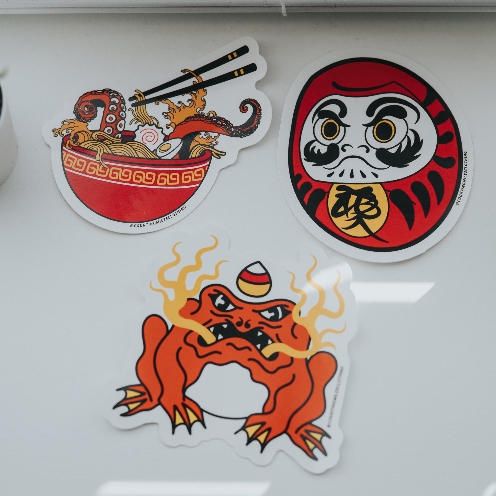 Three decorative stickers on a window, including a bowl, a daruma, and a frog-like creature.