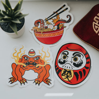 Four colorful stickers on a white surface with a plant and red fabric in the background.