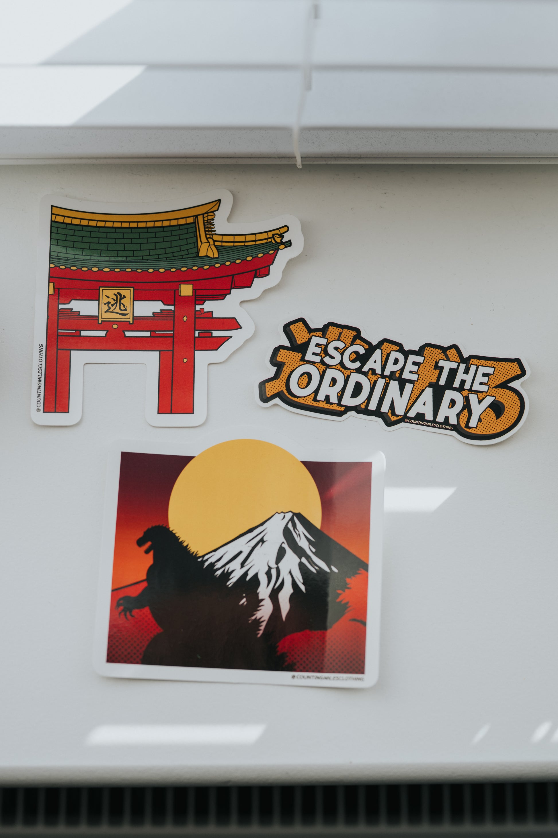 Three stickers on a white surface: a red torii gate, 'Escape the Ordinary' logo, and Godzilla silhouette with Mount Fuji.