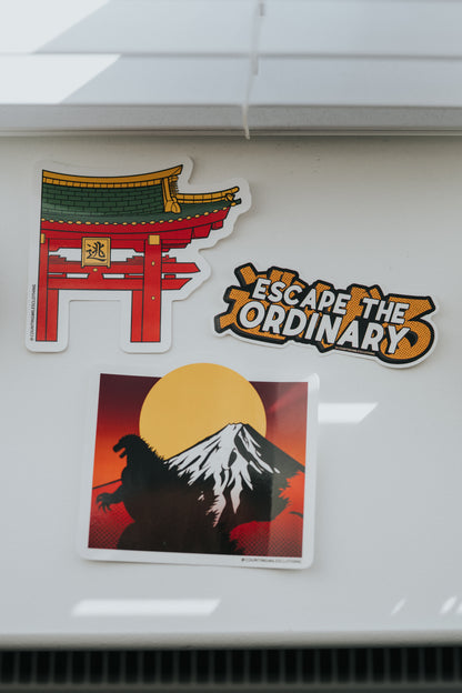 Three stickers on a white surface: a red torii gate, 'Escape the Ordinary' logo, and Godzilla silhouette with Mount Fuji.